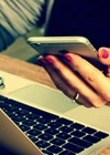 Photo of person sitting at laptop holding mobile phone.